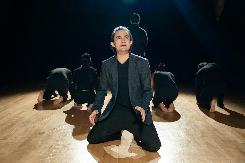 Actor kneels in mid-performance, looking at the audience, while a group of other actors kneel behind him with their backs to the audience.